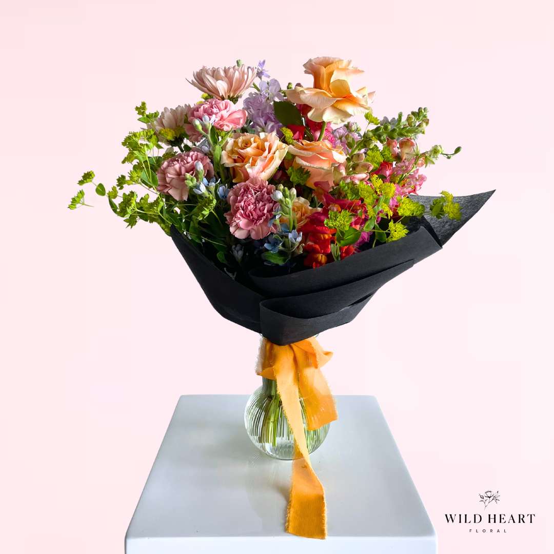 A lush mix of seasonal blooms with various colors in a hand tied bouquet wrapped in waterproof paper.