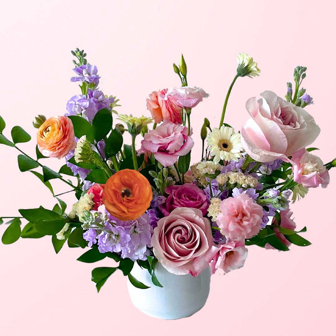 A soft mix of pastel flowers in pinks and lavender hues with pops of orange featuring a mix of stock, ranunculus, roses, lisianthus, accent flowers and garden greens artfully and elegantly designed in a white ceramic vase.