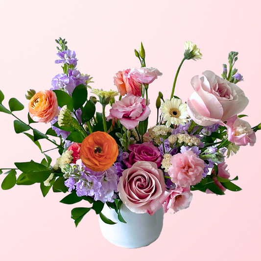 A soft mix of pastel flowers in pinks and lavender hues with pops of orange featuring a mix of stock, ranunculus, roses, lisianthus, accent flowers and garden greens artfully and elegantly designed in a white ceramic vase.