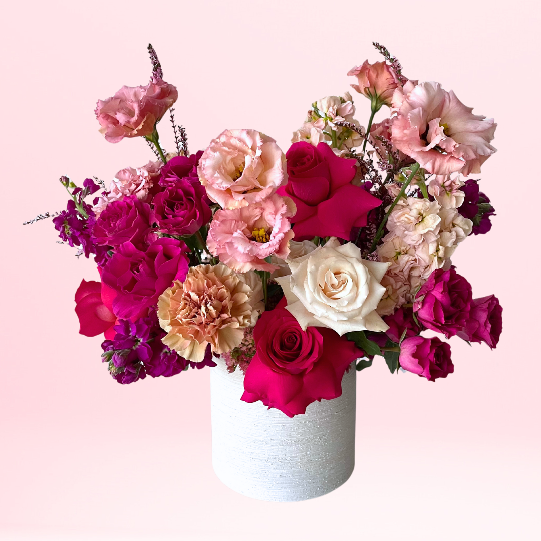 A lush and premium mix of blush, pink, and hot pink flowers featuring a mix of roses, spray roses, lisianthus, stock, and delicate filler, this a floral heavy arrangement with minimal greenery complete with an elegant white ceramic wide cylinder vase.