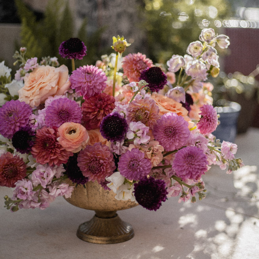 A truly elegant and lush floral arrangement with a mix of pink, lavender, and dark purple and peach seasonal blooms with a floral heavy and artfully designed bouquet housed in a high end gold compote bowl.