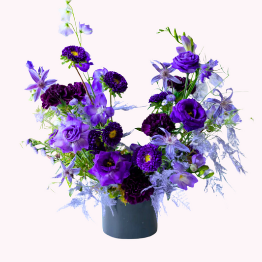 A Fun , vibrant, and modern arrangement with mix of various purple premium flowers and foliage with a modern and artful shape housed in a black ceramic vase.