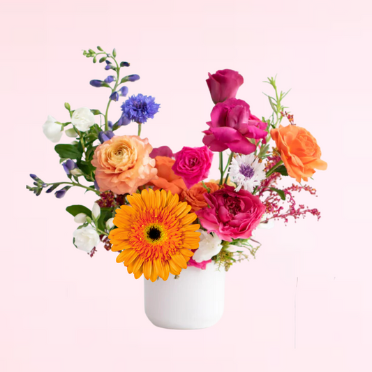 A fun and vibrant artfully designed arrangement with colorful mix of hot pinks, peaches, and oranges, with various pops of elegant filler and other in season blooms. Complete with a classic white ceramic vase.