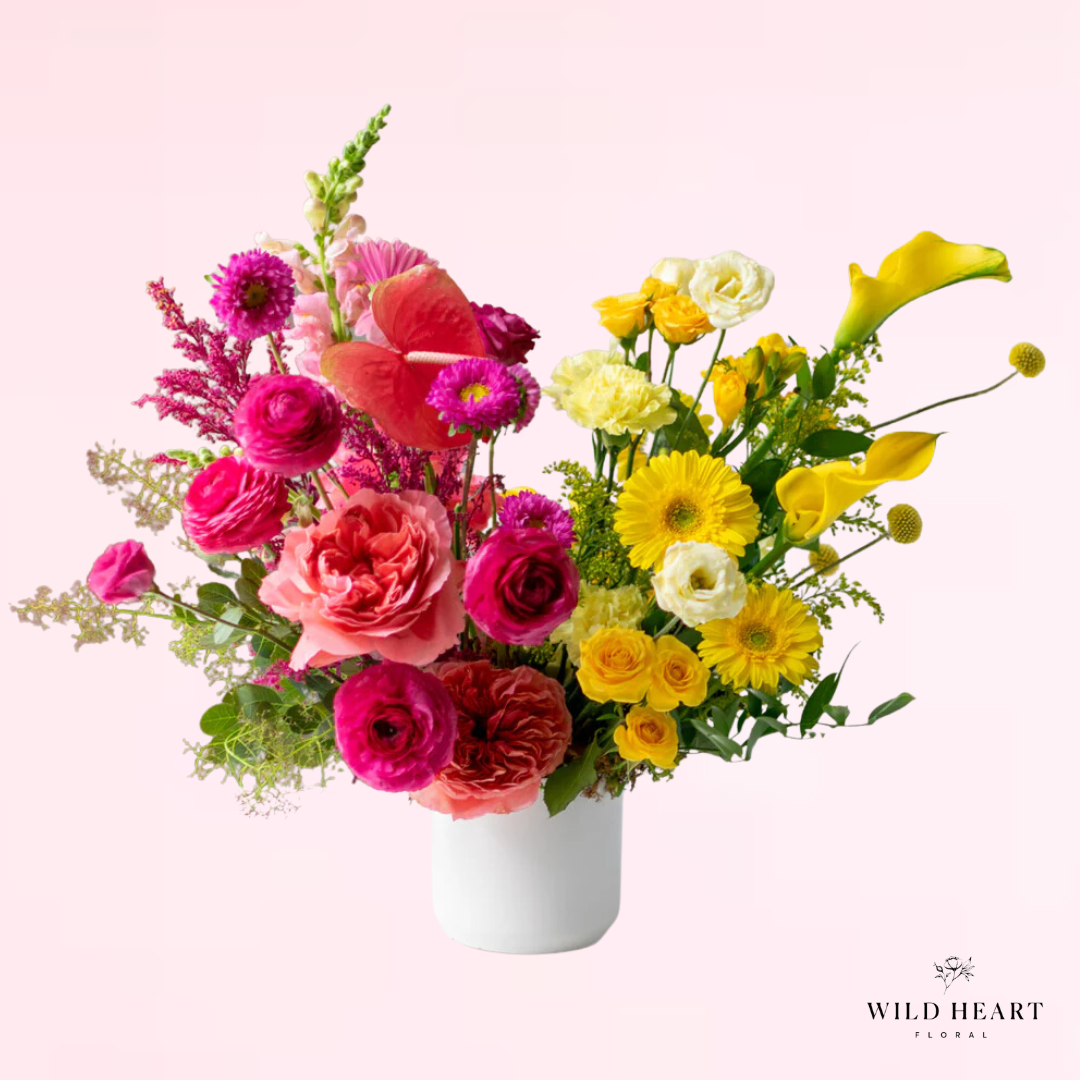 A luscious arrangement featuring vibrant mix of hot pink, pink and yellow premium blooms such as roses, spray roses, gerbera daisies, calla lilies, ranunculus, and snapdragons, with accent blooms and greens designed in a modern and elegant shape.